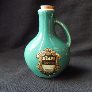 May include: A teal ceramic bottle with a single handle and a cork stopper. The bottle features a decorative black and gold label with the text Dolfi, Strasbourg France, Crème de Menthe, and Product of France printed on the front surface.