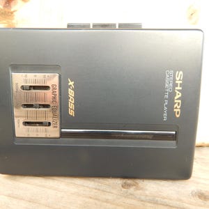 May include: A black Sharp stereo cassette player with a gold X-Bass graphic equalizer. The player has a graphic equalizer with three sliders for 50Hz, 1kHz, and 10kHz.