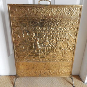 Puede incluir: Una pantalla de chimenea rectangular de latón con una escena en relieve de personas reunidas alrededor de una mesa en una taberna. La pantalla tiene un borde decorativo con motivos florales en la parte superior e inferior y un asa pequeña.