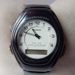 May include: Black Casio digital watch with a white face, featuring analog hands and a digital display. The watch has the text "ILLUMINATOR", "CASIO", "DUAL TIME", and "WATER 100M RESIST". The watch is water resistant.