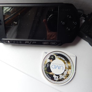 Pode incluir: Uma consola de jogos portátil Sony PSP preta com ecrã panorâmico, painel direcional e botões de ação, acompanhada por um adaptador de corrente preto e um disco Universal Media Disc branco para o jogo Medal of Honor Heroes.