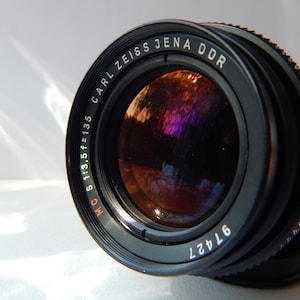 May include: Close-up of a black Carl Zeiss Jena DDR camera lens. The lens has the text "CARL ZEISS JENA DDR" and "MC S 1:3.5 f=135" printed on it. The lens has a reflective interior with a series of ridges around the edge.