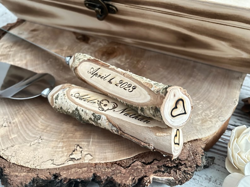 Puede incluir: Un juego de dos cuchillos para cortar pastel de madera con mangos en forma de coraz&oacute;n. Los mangos est&aacute;n grabados con los nombres "Adele & Nathan" y la fecha "April 4, 2023".