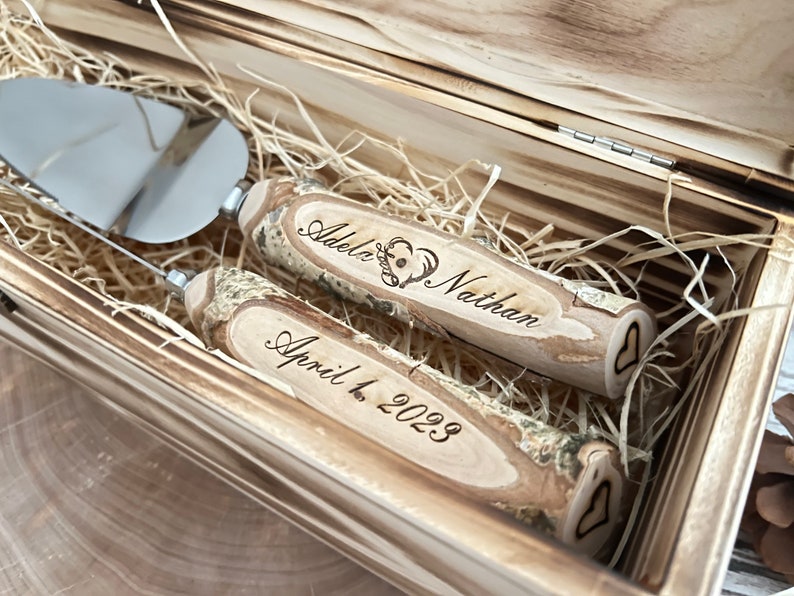 Puede incluir: Una caja de madera con un par de cuchillos para cortar pastel con mangos de madera. Los mangos est&aacute;n grabados con los nombres "Adele & Nathan" y la fecha "April 1, 2023".