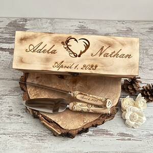 Puede incluir: Una caja de madera grabada con los nombres "Adela" y "Nathan", un dise&ntilde;o de coraz&oacute;n y astas, y la fecha "1 de abril de 2023". La caja est&aacute; abierta, revelando un cuchillo para pastel y un servidor.