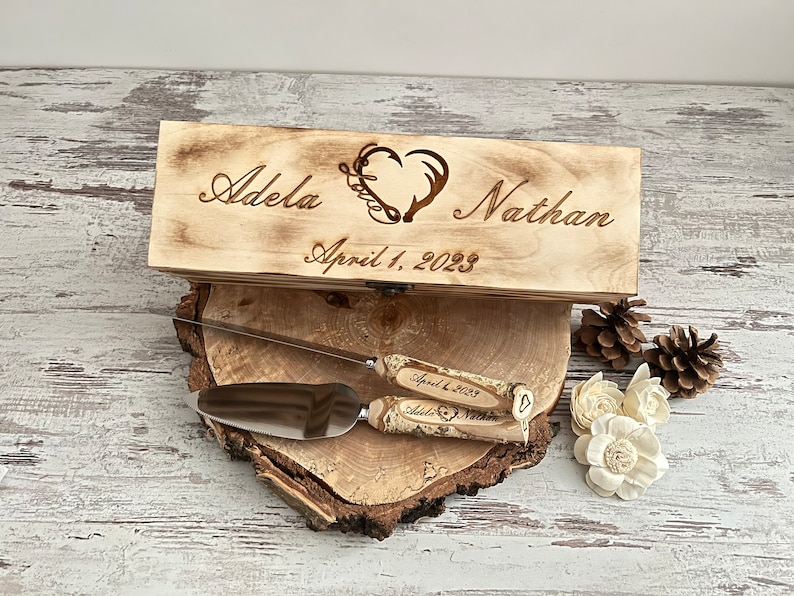 Puede incluir: Una caja de madera grabada con los nombres "Adela" y "Nathan", un dise&ntilde;o de coraz&oacute;n y astas, y la fecha "1 de abril de 2023". La caja est&aacute; abierta, revelando un cuchillo para pastel y un servidor.
