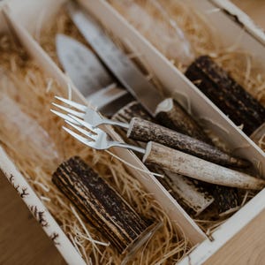 May include: A set of two forks with antler handles in a wooden box with straw. The forks are made of stainless steel and have a polished finish. The antler handles are natural and have a brown color.
