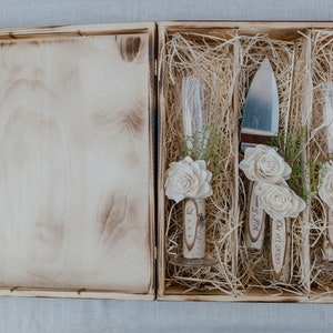 Könnte beinhalten: Eine Holzkiste mit einem Scharnierdeckel, die ein Kuchmesser und zwei Champagnerflöten enthält. Die Flöten sind mit weißen Blumen verziert und das Datum "04.11.2020" ist in das Messer eingraviert.
