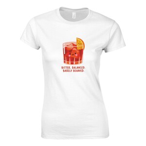 Puede incluir: Una camiseta blanca de manga corta con un gráfico de un cóctel rojo en un vaso con cubitos de hielo, una rodaja de naranja y una cara sonriente. Debajo de la ilustración, el texto dice: BITTER. BALANCED. BARELY BEHAVED. en letras rojas.