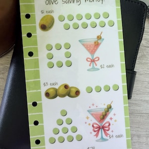 May include: A rectangular light green savings tracker insert with six holes on the left side. It features the text olive saving money!, illustrations of olives and martini glasses, rows of circles for tracking, and the text mandysmonies_budgets.