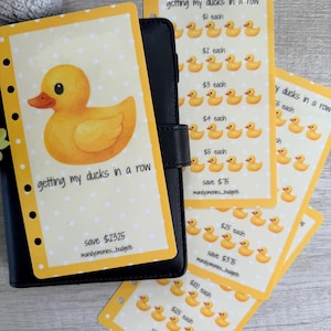 May include: A black planner with yellow and white inserts featuring a cartoon duck and the phrase "getting my ducks in a row." The inserts have a dotted background and a duck graphic. Additional inserts show rows of ducks with dollar amounts.
