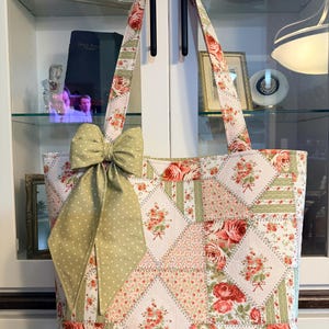 May include: A tote bag featuring a patchwork design of floral patterns in shades of red, green, and cream. The bag has two shoulder straps and is decorated with a large, sage green bow with white polka dots attached to the front left side.