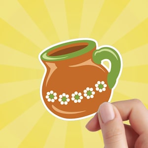 May include: A brown clay pitcher with a green handle and white flower accents. The pitcher is a traditional Mexican design.
