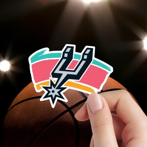 May include: A sticker featuring the San Antonio Spurs logo in black and white, layered over a horizontal brushstroke design in teal, pink, and orange. The sticker is positioned in front of a textured brown basketball with black seams.
