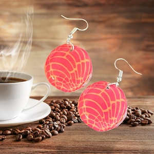 May include: A pair of pink and orange dangle earrings with a geometric pattern. The earrings are hanging from silver hooks.