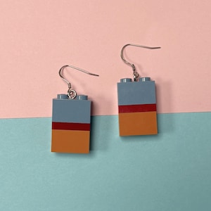 Peut inclure: Une paire de boucles d'oreilles faites à la main avec un design de brique Lego. Chaque boucle d'oreille a une partie supérieure bleue, une bande rouge et une partie inférieure orange. Les boucles d'oreilles sont suspendues à des crochets d'oreille argentés. Le fond est un bloc de couleur rose et bleu.