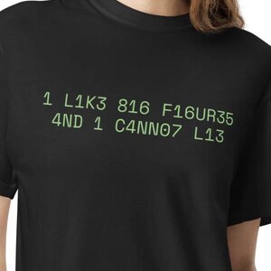 May include: A black short-sleeved t-shirt featuring light green text printed on the chest that reads: 1 L1K3 816 F16UR35 4ND 1 C4NN07 L13. The shirt has a crew neckline and a standard fit, displaying the alphanumeric graphic clearly against the dark fabric.
