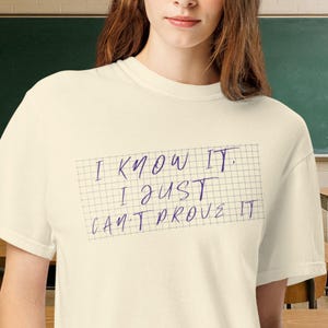 May include: A cream-coloured short-sleeve t-shirt featuring a graphic of a grid pattern with the purple handwritten text "I KNOW IT. I JUST CAN'T PROVE IT" centred on the chest. The shirt has a crew neckline and a standard fit.