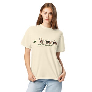 Könnte beinhalten: Cremefarbenes T-Shirt mit einem skurrilen Design. Die Grafik zeigt einen grünen Frosch und eine Reihe von Katzen mit bunten Mustern, begleitet vom Text "are you coming?". Das Shirt besteht aus weichem Material.