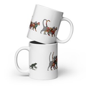 Puede incluir: Dos tazas de cerámica blanca apiladas una encima de la otra. Una taza tiene una silueta de rana y siluetas de gatos con diseños de patchwork coloridos. La otra taza tiene siluetas de gatos con diseños de patchwork coloridos.