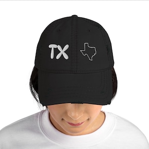 May include: A black baseball cap with a distressed, worn-in finish. The front features white embroidered text that reads TX followed by a white embroidered outline of the state of Texas. The cap has a structured crown and a curved brim.