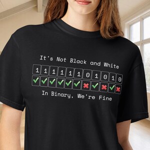 May include: A black short-sleeved t-shirt featuring white text that reads It's Not Black and White, followed by a row of binary digits 1 and 0 in boxes, a row of green checkmarks and red x marks, and the text In Binary, We're Fine at the bottom.