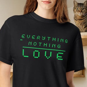 May include: A black short-sleeved t-shirt featuring green dot-matrix style text that reads EVERYTHING - NOTHING, with the word LOVE printed in larger green dot-matrix letters underneath, separated by a horizontal line of green dots.