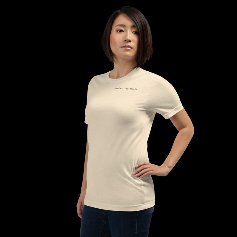 Op de afbeelding: Een cr&egrave;mekleurig T-shirt met de tekst "Readability counts" in een klein, zwart lettertype. Het shirt heeft korte mouwen en lijkt van een zacht materiaal te zijn. De persoon draagt een blauwe spijkerbroek.