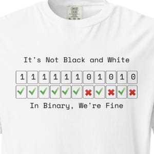 May include: A white t-shirt featuring the text 'It's Not Black and White' above a row of binary digits 1 and 0, each paired with a green checkmark or red x symbol, followed by the text 'In Binary, We're Fine' printed in black ink.