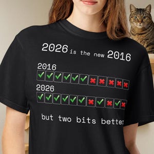 May include: A black t-shirt featuring white text that reads 2026 is the new 2016. Below this, two rows of boxes show checkmarks and red x symbols representing the years 2016 and 2026. The bottom text reads but two bits better in white font.