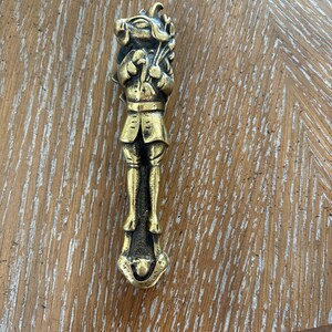 Puede incluir: Un abridor de botellas de latón vintage con forma de figura caprichosa. La figura viste un atuendo detallado y sostiene un paquete de objetos. El abridor tiene un diseño detallado y un cálido color dorado antiguo.