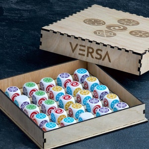 May include: A wooden box with the word "VERSA" printed on the lid, containing 25 white dice with colorful symbols. The box is open and the dice are visible. There are also five cards with colorful symbols on them.