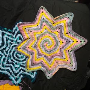 May include: Two handmade crochet star-shaped items. One is a vibrant mix of yellow, pink, and purple with a spiral center. The other is blue and teal with a zigzag pattern. These decorative items are likely used as home decor.