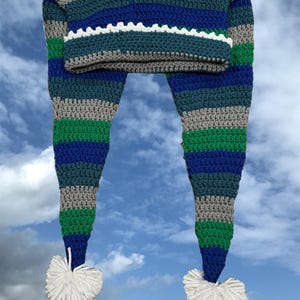 Könnte beinhalten: Eine gestreifte Häkelmütze mit langen Bändern und weißen Bommeln. Die Mütze ist in den Farben Grau, Grün und Blau gehalten.