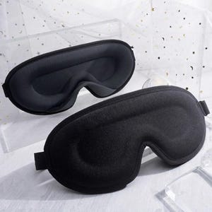 Op de afbeelding: Twee zwarte, voorgevormde slaapmaskers met een gegoten ontwerp, diepe oogkassen en een glad, gewatteerd oppervlak. Elk masker is voorzien van een verstelbare band aan de zijkant, ontworpen om licht te blokkeren en een comfortabele pasvorm te bieden.