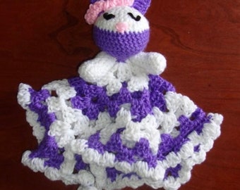 Puede incluir: Una manta de seguridad de conejito de ganchillo hecha a mano en blanco, morado y rosa. El conejito tiene una cara y orejas moradas, un lazo rosa y ojos negros. La manta tiene un patrón de cuadros y el texto "Little Barefoot Life Boutique".