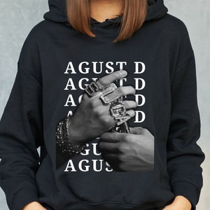 Puede incluir: Sudadera negra con una imagen en blanco y negro de manos adornadas con anillos y pulseras. La palabra "AGUST D" est&aacute; impresa en blanco verticalmente. La sudadera tiene una capucha con cord&oacute;n y un bolsillo de canguro.