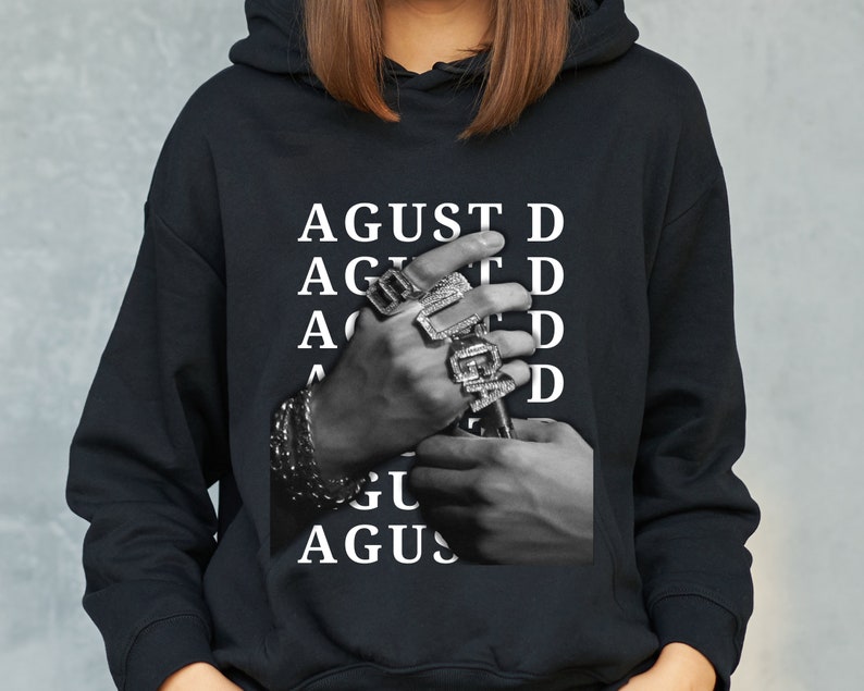 Puede incluir: Sudadera negra con una imagen en blanco y negro de manos adornadas con anillos y pulseras. La palabra "AGUST D" est&aacute; impresa en blanco verticalmente. La sudadera tiene una capucha con cord&oacute;n y un bolsillo de canguro.