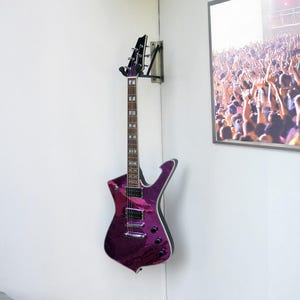 Könnte beinhalten: Eine violette E-Gitarre an einer Wandhalterung. Die Gitarre hat eine einzigartige Körperform und eine dunkle Kopfplatte. Ein gerahmtes Bild einer Konzertmenge ist zu sehen. Die Gitarre wird in einer Ecke des Raumes ausgestellt.