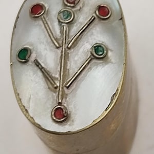 May include: A vintage, oval-shaped jewellery piece with a mother-of-pearl surface. The design features a silver-coloured tree-like pattern with red and green gemstone accents. The base is a gold-coloured metal.