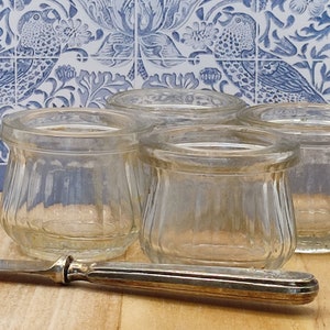 May include: Four small, clear glass jars with a ribbed design, arranged on a wooden surface. A silver knife lies in front of the jars. The background features a blue and white floral tile pattern.
