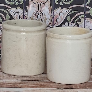May include: Two cylindrical off-white ceramic crocks with a smooth finish and a slightly raised rim. The vessels are plain, featuring a neutral cream color and a simple, straight-sided design. These containers are uniform in shape and size.
