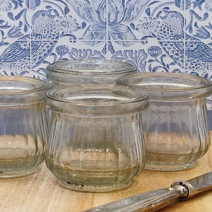 May include: Four small, clear glass jars with a ribbed design, arranged on a wooden surface. A silver knife rests nearby. The background features blue and white patterned tiles with a bird motif. The jars are empty.