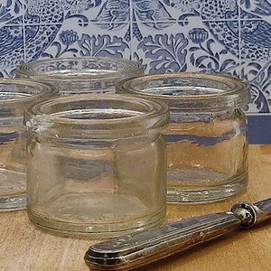 May include: Four clear glass jars with a silver butter knife on a wooden surface. The jars are round and have a wide mouth.
