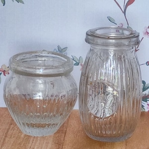 May include: Two clear glass jars with textured surfaces. The jar on the left has a rounded shape, while the jar on the right has a taller, more cylindrical shape.