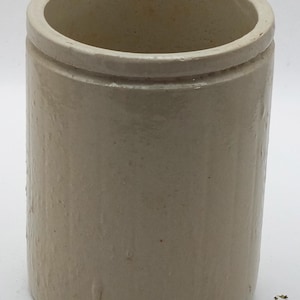 May include: A cylindrical off-white stoneware crock with a smooth, glazed finish and a slightly raised rim around the top opening. The vessel features a simple, vertical structure with a uniform color and a subtle, textured surface throughout.