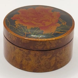 May include: A round lidded box with a glossy finish. The lid features a large orange and red flower design on a black background. The sides of the box and the lid edge have a mottled brown and gold pattern with a smooth, reflective surface.
