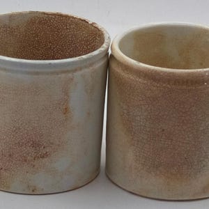 May include: Two cylindrical ceramic crocks featuring a cream-colored base with a distinct brown crackle glaze finish. Both vessels have a smooth, straight-sided profile and an open top, displaying a weathered, aged appearance with visible surface texture.