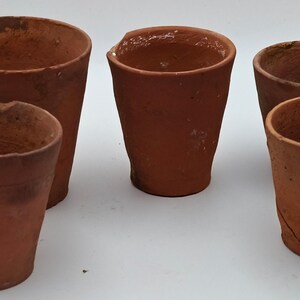 May include: A collection of four terracotta pots with a rustic, unglazed finish. These vessels feature varying sizes, cylindrical shapes, and weathered textures with visible surface imperfections, chips, and earthy brown tones throughout.