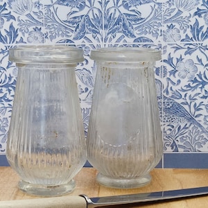 May include: Two clear glass jars with ribbed sides. The jars are empty and have a slightly cloudy appearance. The jars are sitting on a wooden surface.